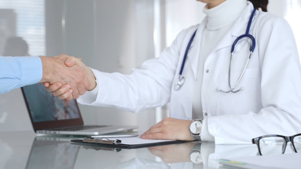Hands of medical professional and male shaking across a desk, symbolizing trust and partnership.